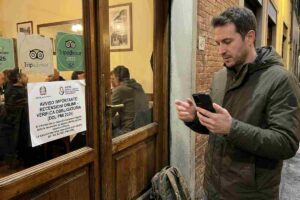 Un uomo con lo smartphone guarda un cartello sulla porta di un ristorante italiano che annuncia: "DDL PMI 2026 - Verifica Recensioni Online Obbligatoria". La scena, scattata in strada a Firenze di sera, mostra l'impatto reale della nuova legge anti-recensioni false.